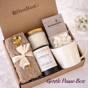 Puede incluir: Una caja de regalo BlissBox marr&oacute;n con una vela, t&eacute;, taza, cuchara y una manta suave atada con una cinta. La caja incluye un paquete de t&eacute; de Moonstruck y malvaviscos. Las palabras "Tea with Tae" son visibles en un recipiente de t&eacute;.