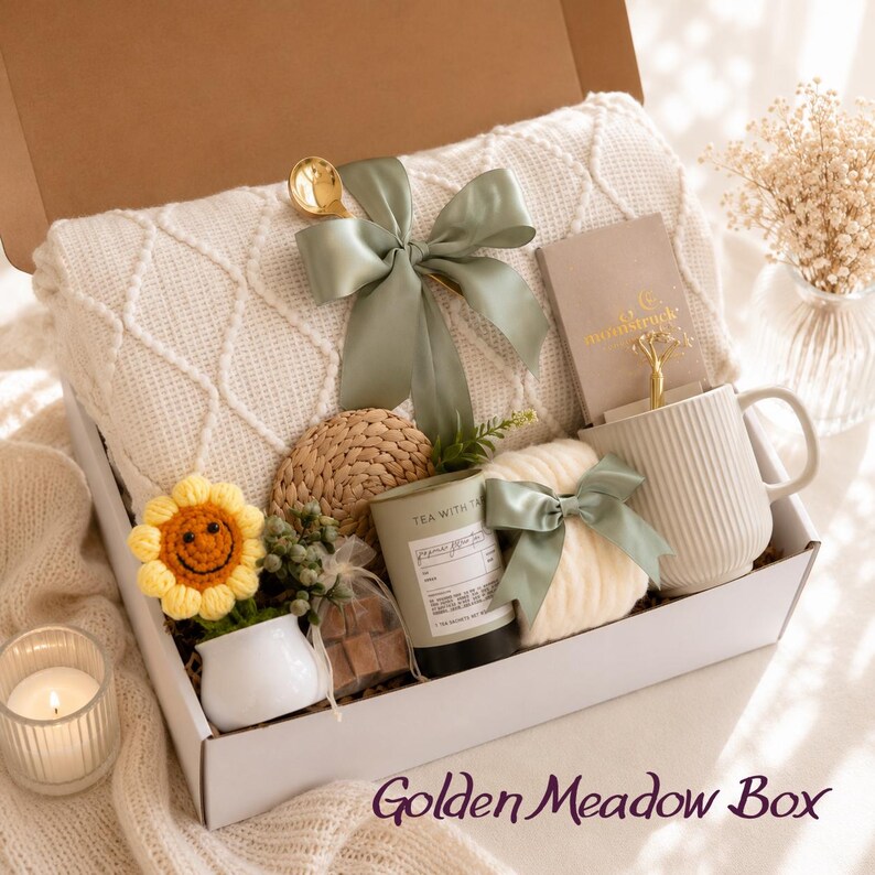 May include: A gift box labeled "Golden Meadow Box" filled with a cream-colored knitted blanket, a mug, a candle, a small sunflower, and other items. The box is decorated with sage green bows and a gold spoon.