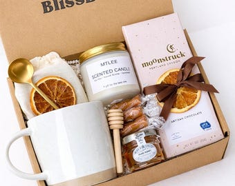 May include: A gift box labeled "Bliss Box" filled with a white mug, candle, chocolate bar, honey, caramels, and a gold spoon. The candle is labeled "MTLEE Scented Candle". The box is labeled "Caramel Delight Box".