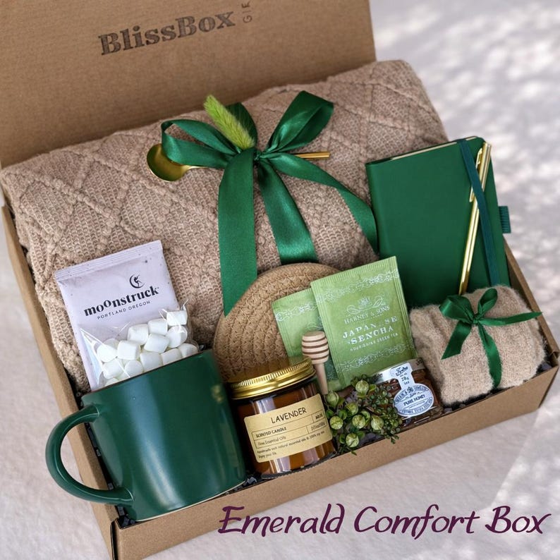 May include: A gift box labeled BlissBox containing a beige cable-knit blanket tied with a green ribbon, a dark green mug, a bag of marshmallows, a lavender candle, a green notebook with a gold pen, a packet of Japan Sencha tea, and honey.