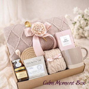 May include: A gift box filled with self-care items. Includes a beige blanket, mug, hot cocoa, tea, honey, and a decorative flower. The box is tied with a pink ribbon and has the text "Calm Moment Box".