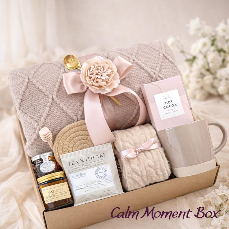 May include: A gift box filled with self-care items. Includes a beige blanket, mug, hot cocoa, tea, honey, and a decorative flower. The box is tied with a pink ribbon and has the text "Calm Moment Box".