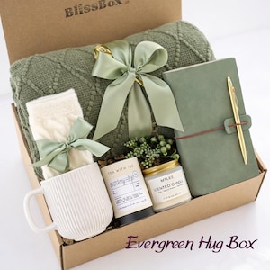 May include: An Evergreen Hug Box filled with a green cable-knit blanket, a journal with a gold pen, a white mug, socks, a candle, and tea. The box is tied with a green ribbon. The box is in a cardboard box.