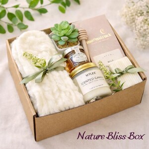 May include: A gift box labeled "Nature Bliss Box" filled with a variety of items. Includes a white fluffy pair of mittens, a small succulent plant, a jar of honey, a scented candle, and a small notebook. The box is tied with green ribbon.