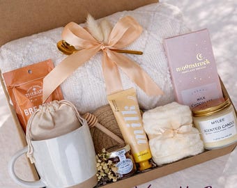 May include: A gift box containing a white cable-knit blanket tied with a peach ribbon, a ceramic mug, a pouch, a honey dipper, a jar of honey, a tube of milk lotion, a bar of Moonstruck milk chocolate, and a scented candle by Mtlee.