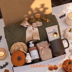 Könnte beinhalten: Eine Geschenkbox mit einer olivgrünen Strickdecke mit beigem Band, einer Moonstruck-Schokolade, einem braunen gerippten Becher mit goldenem Löffel, einem beigen Strickartikel, einem Teeglas, einem geflochtenen Untersetzer, einem kleinen orangefarbenen Kürbis und einer Geisterfigur.