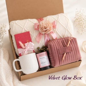 May include: A gift box containing a white cable-knit blanket with a pink ribbon and flower, a white ribbed mug, a red chocolate bar with a pink bow, a black tea tin, and a mauve velvet zippered pouch with a gold pen. Text reads Velvet Glow Box.