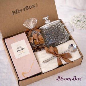 May include: A gift box with a clear glass mug with a bear-shaped lid, a pink box of hot cocoa, a bag of caramel candies, a small spoon, and a white fluffy cloth, all tied with brown ribbons. The box is brown and has the text "BlissBox" and "Bloom Box".