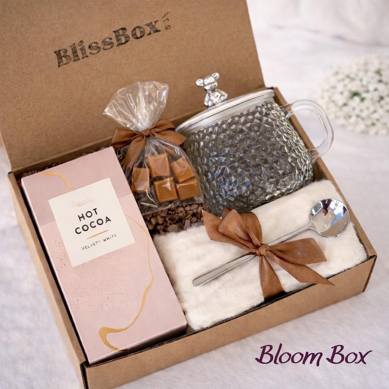 May include: A gift box with a clear glass mug with a bear-shaped lid, a pink box of hot cocoa, a bag of caramel candies, a small spoon, and a white fluffy cloth, all tied with brown ribbons. The box is brown and has the text "BlissBox" and "Bloom Box".