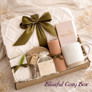 May include: A gift box filled with cozy items. Includes a white knitted blanket with a green bow, a candle, a mug, a coaster, a soap bar, and a tea tube. The box is labeled "Blissful Cozy Box".