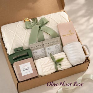 Op de afbeelding: Een geschenkdoos met een witte kabelgebreide deken vastgebonden met een saliegroen lint, een Moonstruck chocoladereep, een witte geribbelde keramische mok, twee Tea With Tae theezakjes, een groene haarklem en een doosje Sea Salt Caramel koekjes.