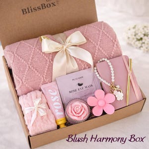 May include: A gift box labeled BlissBox containing a pink diamond-patterned knit blanket tied with a cream ribbon, a rose eye mask packet, a rose-shaped candle, a pink notebook with a gold pen, a pearl bracelet, and pink knit socks.