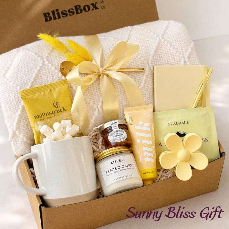 May include: A gift box filled with self-care items. Includes a white mug, a candle, honey, a face mask, and a cream-colored blanket tied with a yellow ribbon. The box is labeled "BlissBox" and "Sunny Bliss Gift".