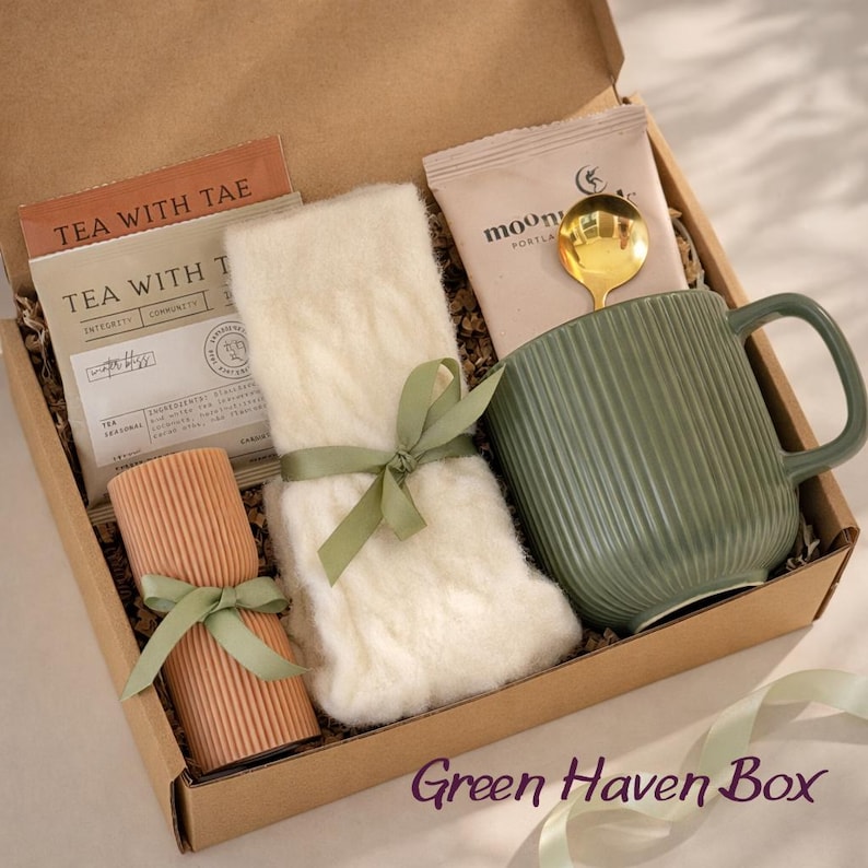 Op de afbeelding: Een geschenkdoos met een Tea with Tae theezakje, een paar witte kabelgebreide sokken met een groen lint, een beige geribbelde kaars met een groen lint, een Mooni Portland zakje met een gouden lepel en een saliegroene geribbelde mok.