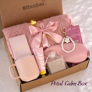 Op de afbeelding: Een BlissBox geschenkdoos met een roze textuurdeken met satijnen strik, een gouden lepel, een roze mok, rozenhandcr&egrave;me, een stuk zeep in een organza zakje, een roze slaapmasker, een parel-sleutelhanger en een Moonstruck-kaart in roze verpakking.