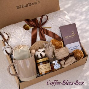 May include: A brown cardboard gift box labeled "BlissBox" contains a white blanket, a mug, a candle, a chocolate bar, and other items. The box is tied with a brown ribbon and includes the text "Coffee Bliss Box".