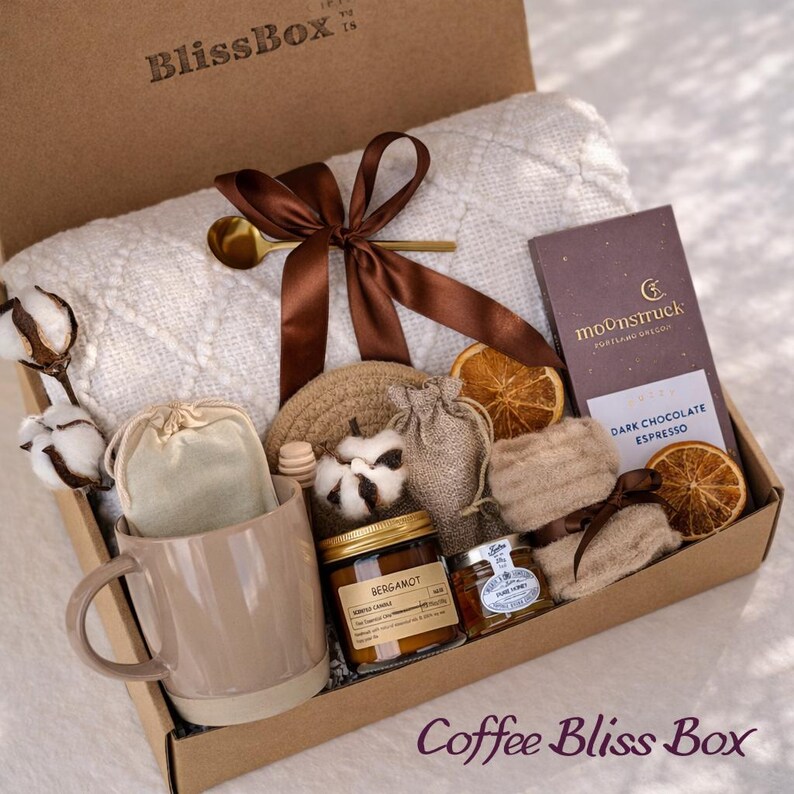 May include: A brown cardboard gift box labeled "BlissBox" contains a white blanket, a mug, a candle, a chocolate bar, and other items. The box is tied with a brown ribbon and includes the text "Coffee Bliss Box".