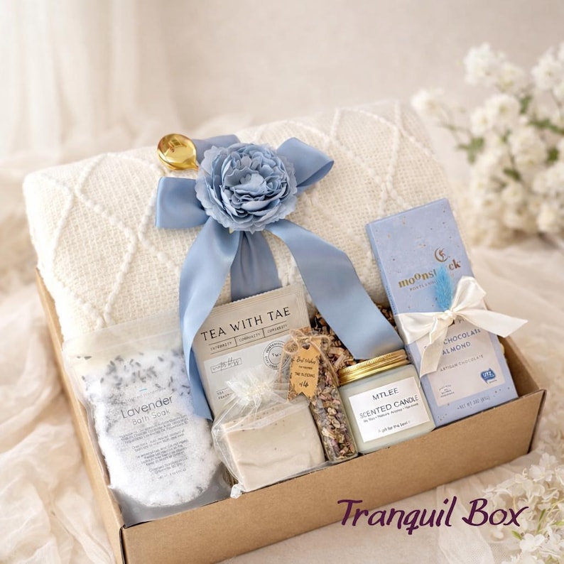 May include: A gift box with a cream-colored blanket, lavender bath soak, tea, soap, a scented candle, and chocolate. The box is tied with a light blue ribbon and flower. The text "Tranquil Box" is visible.