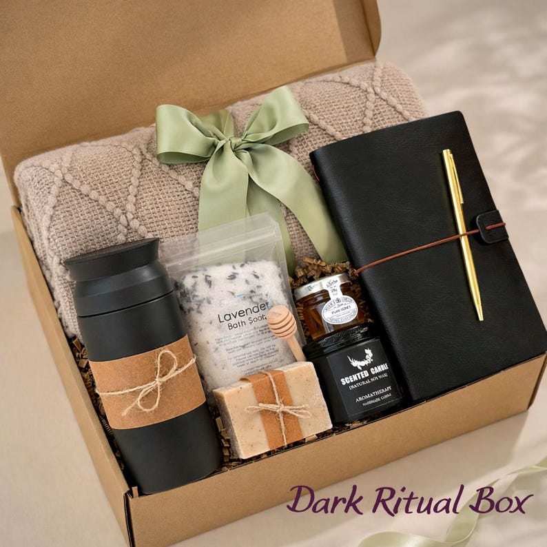 May include: A gift box containing a beige knit blanket with a sage green ribbon, a black travel mug with a brown paper wrap, a pouch of lavender bath soak, a small honey jar with a wooden dipper, a bar of soap, and a black journal with a gold pen.