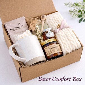 May include: A cardboard gift box containing a white ceramic mug, a bag of marshmallows labeled Moonstruck, a glass vial of dried flowers, a wooden honey dipper, a small jar of honey, a strawberry scented candle, and a cream cable knit scarf.