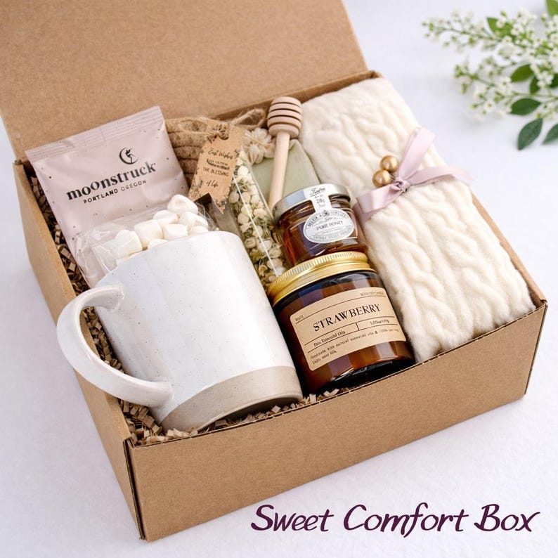 May include: A cardboard gift box containing a white ceramic mug, a bag of marshmallows labeled Moonstruck, a glass vial of dried flowers, a wooden honey dipper, a small jar of honey, a strawberry scented candle, and a cream cable knit scarf.