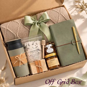 May include: A gift box containing a beige cable-knit blanket with a sage green ribbon, a sage green tumbler with a brown band, a pouch of lavender bath soak, a wooden honey dipper, a bar of soap, a jar of honey, a candle, and a sage green journal with a gold pen.