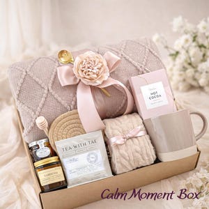 May include: A gift box filled with self-care items. Includes a beige blanket, mug, hot cocoa, tea, honey, and a small knitted item, all tied with a pink ribbon. The box is labeled "Calm Moment Box".