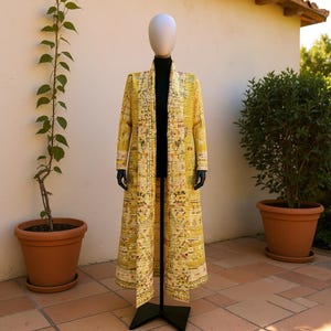 May include: A long, yellow duster coat with a unique pattern, displayed on a mannequin. The coat features a mix of yellow, cream, and red hues. The mannequin is on a black stand, set against a neutral background with potted plants.