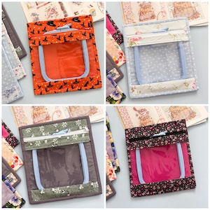 May include: A collection of four square fabric pouches, each featuring a clear plastic window and a zippered top compartment. The pouches display various patterns including orange with black witch hats, light blue polka dots, green floral, and black floral.