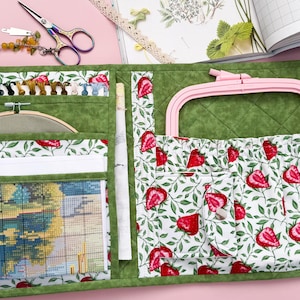 May include: A quilted fabric craft organizer in green with a strawberry and leaf print. The interior features multiple pockets, a clear vinyl sleeve containing a cross-stitch pattern, a wooden embroidery hoop, and a rectangular pink plastic frame.