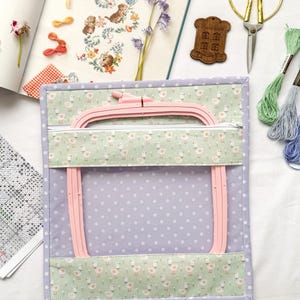Puede incluir: Un organizador de manualidades con un bastidor de bordado rosa, con un estampado de lunares lavanda y patos. El organizador tiene un bolsillo con cremallera y está rodeado de suministros de manualidades, incluyendo tijeras, hilo y un patrón de punto de cruz.