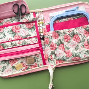 May include: A pink floral print craft organizer, open to display its contents. It features a variety of pockets, zippers, and compartments holding scissors, embroidery floss, and a blue embroidery hoop. The fabric has a rose pattern.