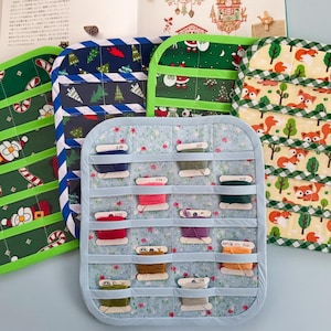Könnte beinhalten: Fünf rechteckige Stoff-Organizer mit horizontalen Taschen für Stickgarnspulen. Die Organizer zeigen verschiedene Muster wie Weihnachtsmänner, Tannenbäume und Füchse, mit farbiger Stoffeinfassung an den Rändern.