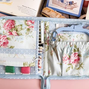 Puede incluir: Kit de costura abierto, con estampado floral azul claro y cierre de cremallera. El kit contiene un bastidor de bordado azul, tijeras, carretes de hilo y un organizador de agujas. La tela interior presenta un patrón de rosas rosas y blancas.