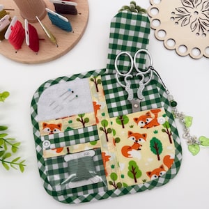 May include: A fabric sewing organiser featuring a green and white gingham pattern with a fox and tree print. It includes a grey felt needle pad, a clear plastic pocket holding a spool of green thread, and a slot containing silver metal scissors.