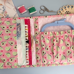 May include: Open craft organiser with a pink avocado print. The organiser holds a blue embroidery hoop, needles, thread, and scissors. The background includes spools of pink and green thread, and a wooden thread organiser.