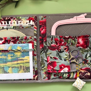 May include: A quilted fabric organizer for embroidery supplies featuring a pomegranate and floral print in red, green, and grey. It includes multiple pockets holding a cross-stitch pattern, a thread organizer, a pink embroidery hoop, and scissors.