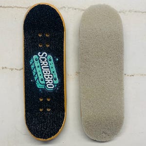 Puede incluir: Un fingerboard negro con el texto "SCRUBBRO" en turquesa y blanco, junto a una cinta de agarre de espuma gris. El fingerboard tiene un borde amarillo y cuatro agujeros para los ejes.