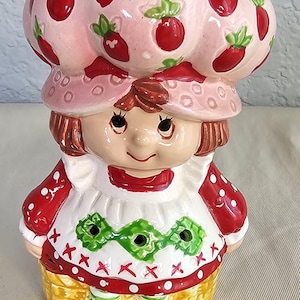 May include: A ceramic figurine of a character wearing a large pink hat decorated with red strawberries and green leaves. The figure has brown hair, a white apron with green diamond patterns and red cross-stitch details, red sleeves, and brown shoes.