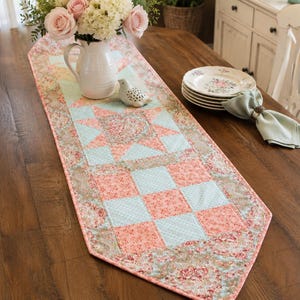May include: A quilted table runner with a patchwork design in shades of coral, light blue, and floral patterns. The runner features a star and checkerboard pattern, adding a touch of country charm to the wooden table. A vase of flowers and plates are also on the table.