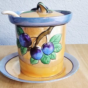 May include: A ceramic jam pot with a matching lid and saucer, featuring a hand-painted design of dark purple plums and green leaves on a golden-yellow background. The rim of the pot, lid, and saucer are finished in a glossy iridescent blue.