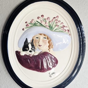 May include: An oval ceramic wall plaque featuring a relief sculpture of a person wearing a wide-brimmed light blue hat adorned with pink flowers, holding a black and white Boston Terrier dog, dressed in a deep purple garment, signed Joni.