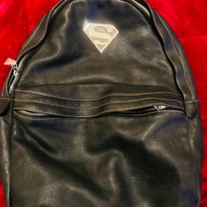 Puede incluir: Mochila de cuero negro con un logotipo de Superman plateado en la parte delantera.