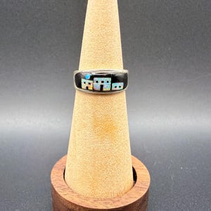May include: A silver ring with a black band featuring a colorful inlaid design of buildings. The ring has a unique, artistic style, and is displayed on a wooden ring holder. The ring's design includes white, blue, and orange colors.