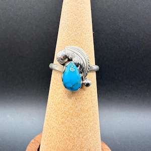 May include: A silver ring featuring a teardrop-shaped turquoise stone, accented by a detailed feather design. The ring is displayed on a wooden stand against a dark background, showcasing the vibrant blue of the stone.