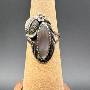 May include: A silver ring featuring an oval, iridescent stone. The ring has a floral accent and a feather design on the side. The ring is displayed on a wooden stand against a dark background.