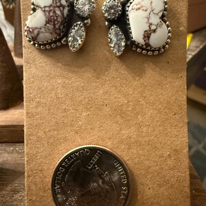 May include: A pair of silver-toned earrings with a white and brown marbled stone design. Each earring features a heart-shaped stone, surrounded by a beaded edge, and accented with clear, round and teardrop-shaped crystals. The earrings are displayed on a brown card.