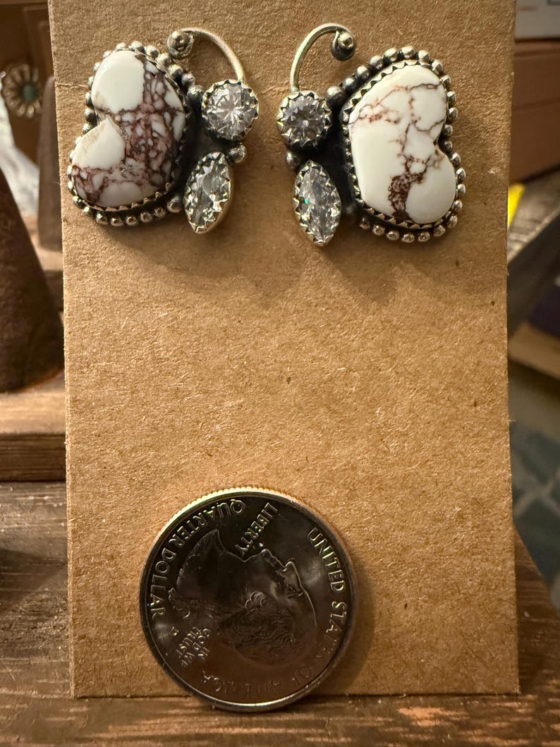 May include: A pair of silver-toned earrings with a white and brown marbled stone design. Each earring features a heart-shaped stone, surrounded by a beaded edge, and accented with clear, round and teardrop-shaped crystals. The earrings are displayed on a brown card.