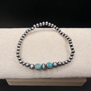 May include: A silver beaded bracelet featuring three turquoise stones. The bracelet is composed of small, round silver beads. Two turquoise stones flank a silver bead. The bracelet is presented on a cream-coloured surface.