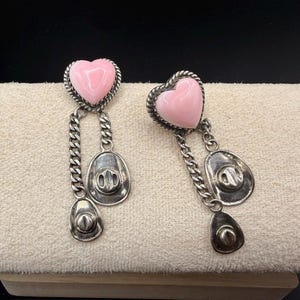 May include: A pair of dangle earrings featuring a smooth, light pink heart-shaped stone set in a twisted silver-tone metal frame. Each earring has two silver-tone metal cowboy hat charms suspended from the heart by short, delicate curb chains.